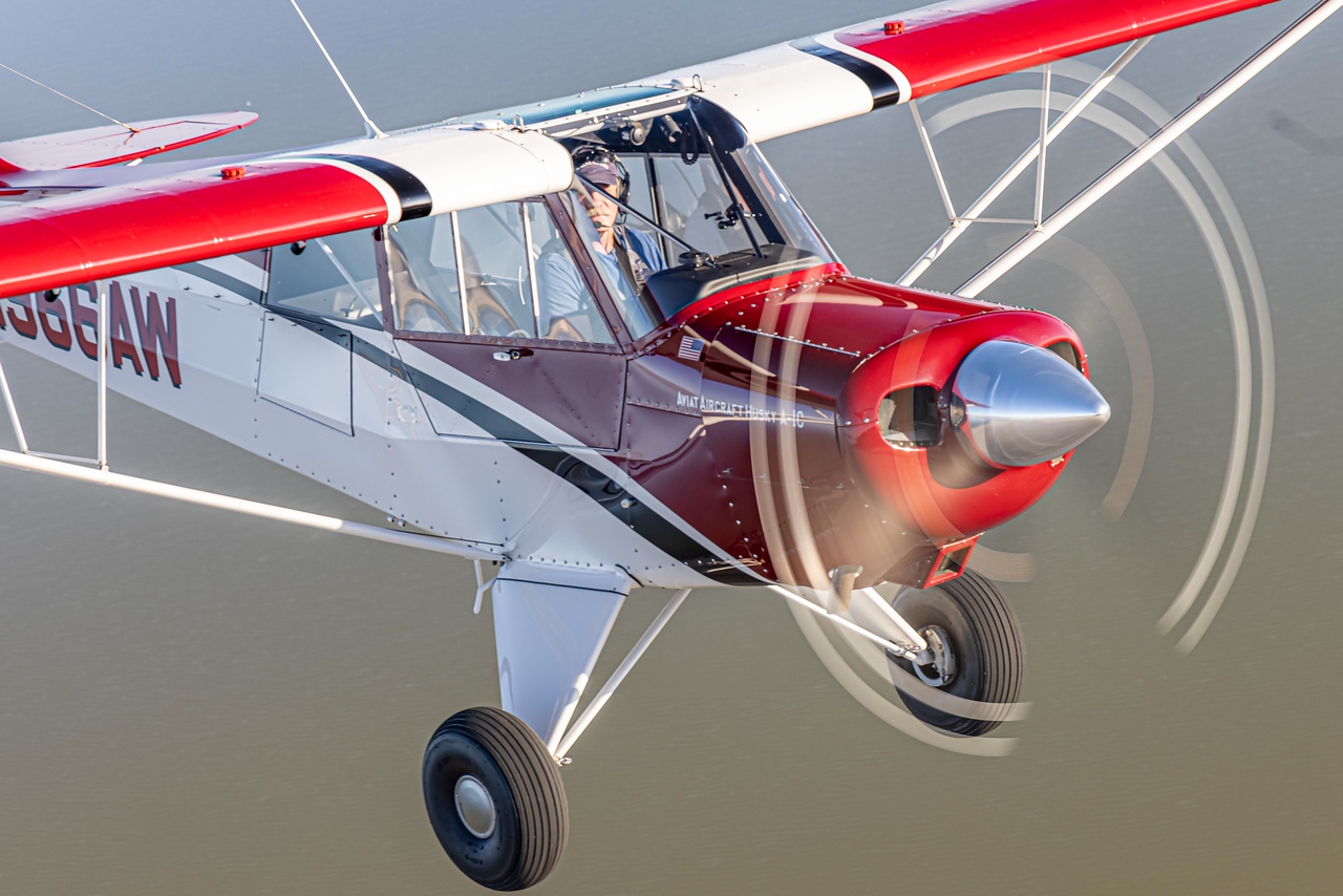 A close view of a red-and-white Husky in flight, showing the cockpit, wing struts and propeller.